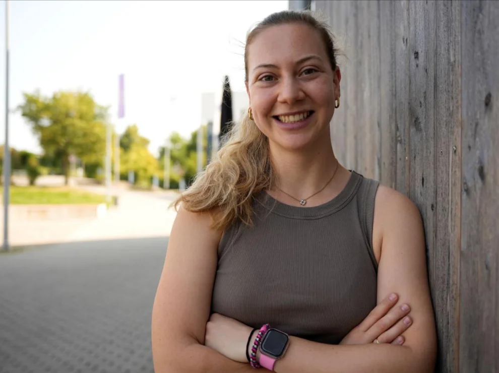 Porträt einer jungen Frau, die mit verschränkten Armen an einer Holzwand lehnt.