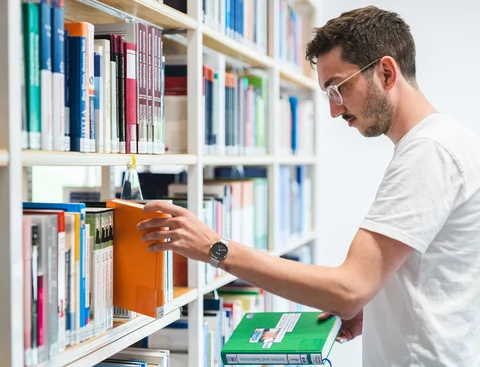 Universitätsbibliothek (Foto: UW/H | Johannes Buldmann) Ein Student steht vor einem Bücherregal und greift nach einem Buch.
