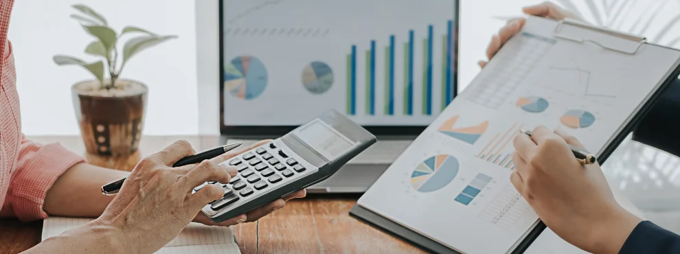 Zwei Menschen sitzen an einem Tisch, eine weibliche Hand hält einen Taschenrechner. Eine männliche Hand zeigt auf Grafiken.