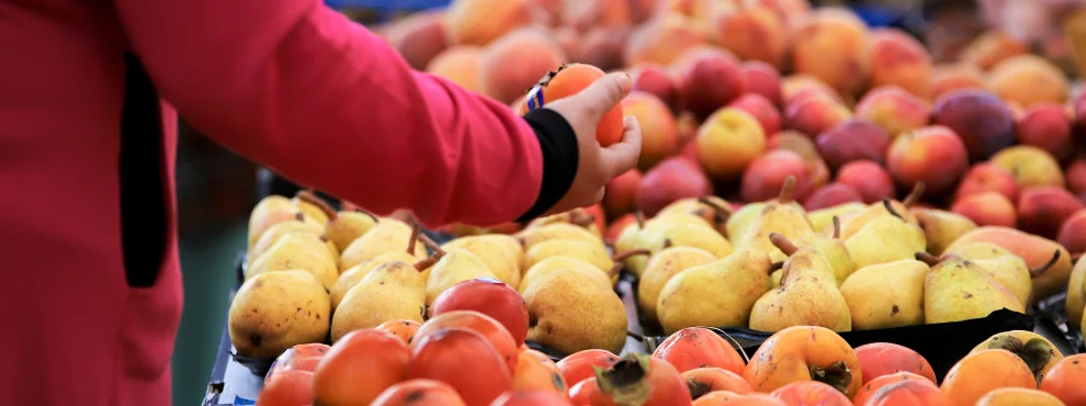 One hand holds a khaki, a large fruit display can be seen in the background.