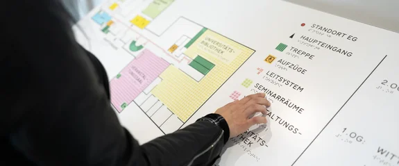 Übersichtsplan des Holzgebäudes (Foto: UW/H | Michael Schwettmann) Eine Person zeigt mit ihrem Finger auf einen taktilen Gebäude-Lageplan.