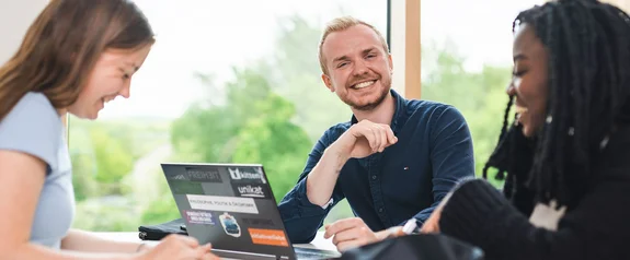 Studierende an der Universität Witten/Herdecke (Foto: UW/H | Johannes Buldmann) Gruppe von drei Studierenden sitzt am Tisch mit Laptop in fröhlicher Stimmung.