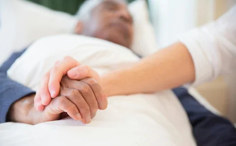 Eine pflegende Person hält die Hand eines alten Mannes, der in einem Bett liegt.