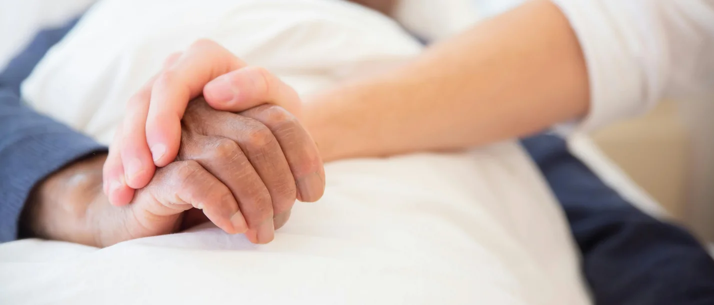 Eine pflegende Person hält die Hand eines alten Mannes, der in einem Bett liegt.