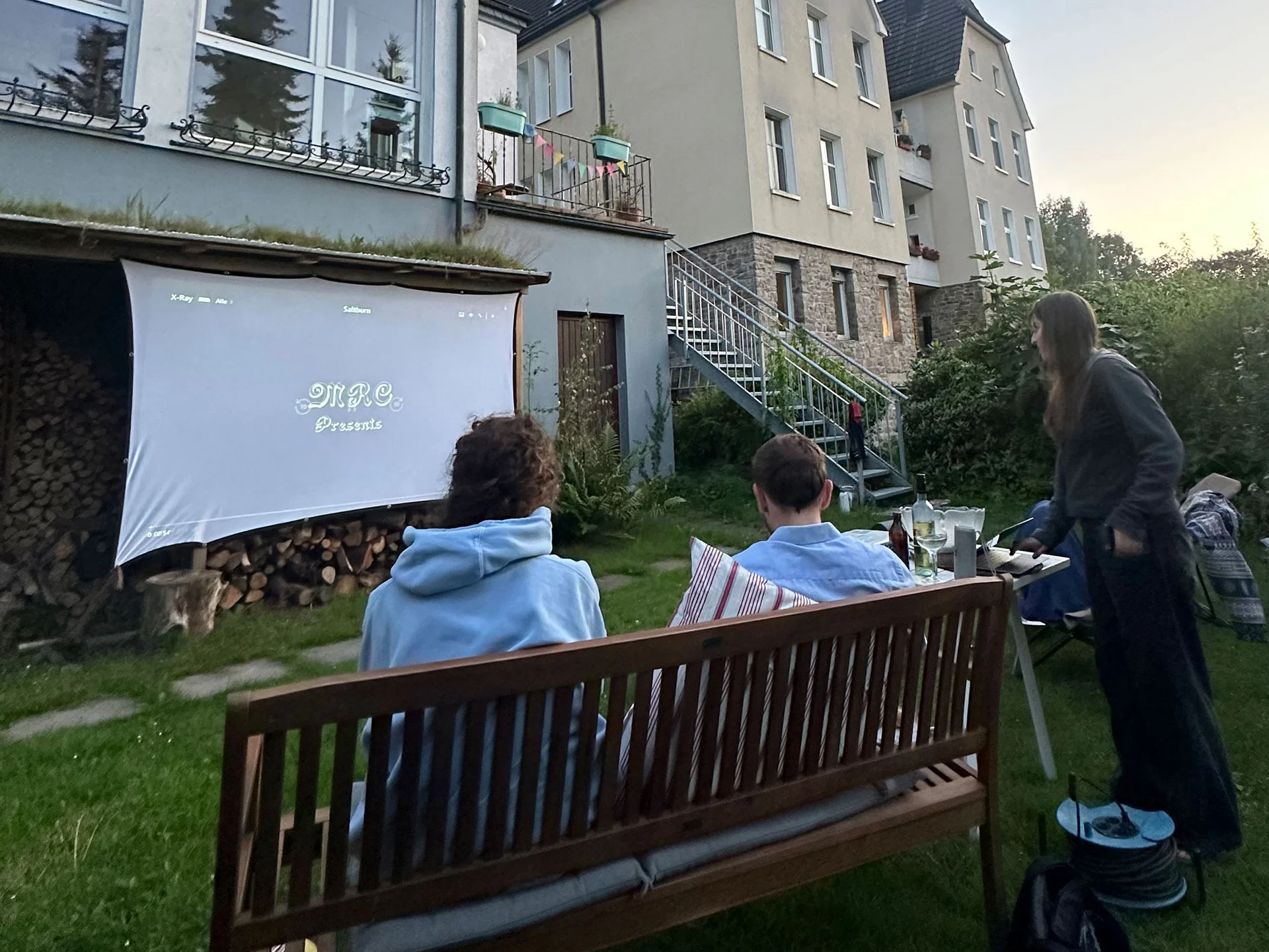Eine Gruppe junger Menschen sitzt auf Bänken und Gartenstühlen auf einer Wiese. Sie schauen auf eine Leinwand, auf die ein Film projiziert wird. Im Hintergrund sind Häuserfassaden.