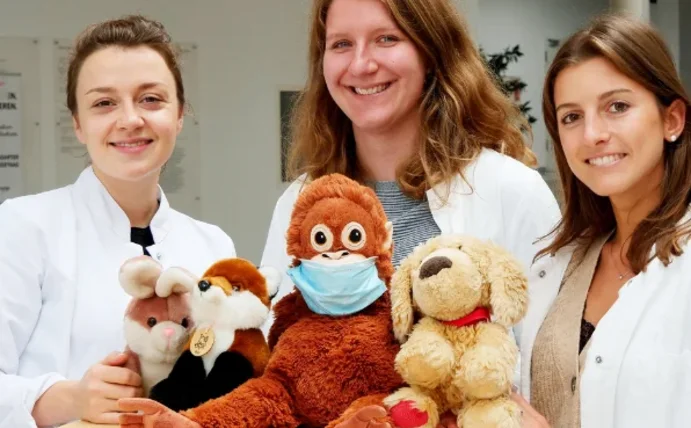 Drei Studentinnen lächeln in die Kamera. Auf einem Tisch vor ihnen befinden sich mehrere Stofftiere.
