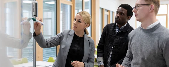 Lerne unsere Studiengänge aus dem Bereich Wirtschaft & Gesellschaft kennen als Studentin für einen Tag. (Foto: UW/H | Michael Schwettmann) Eine Frau erklärt zwei jungen Männern etwas an einem Whiteboard.