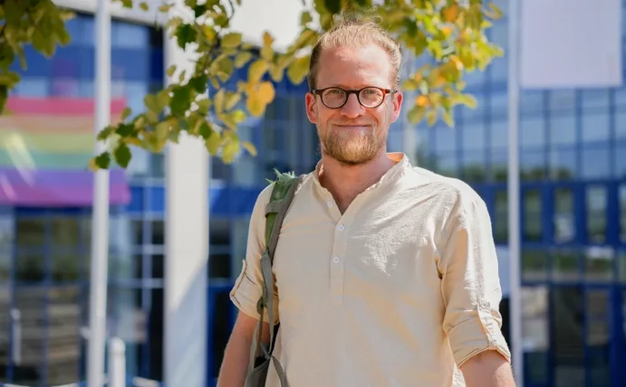 Porträt eines Mannes vor dem Universitätsgebäude. Über der Schulter trägt er einen Rucksack.