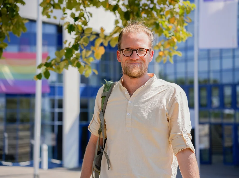 Porträt eines Mannes vor dem Universitätsgebäude. Über der Schulter trägt er einen Rucksack.
