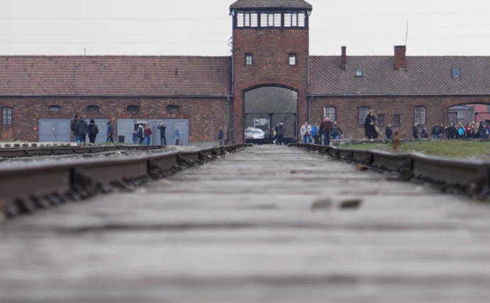 Im Vordergrund sieht man verschwommene Bahnschienen. Im Hintergrund das KZ Auschwitz, drumherum ganz klein ein paar Menschen.