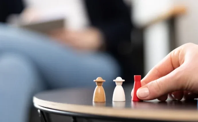 Man sieht eine Therapiesituation. Im Hintergrund eine verschwommene Therapeutin, im Vordergrund die Hand einer Patientin und Spielfiguren.