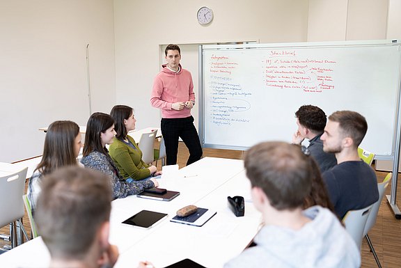 Die Wissenschaftler:innen der UW/H arbeiten in interdisziplinären Forschungsprojekten fakultätsübergreifenden zusammen (Foto: UW/H | Michael Schwettmann) Ein junger Mann steht an einem Whiteboard und schaut in die Gesichter junger Menschen, die im Kreis an einem Tisch vor ihm sitzen.