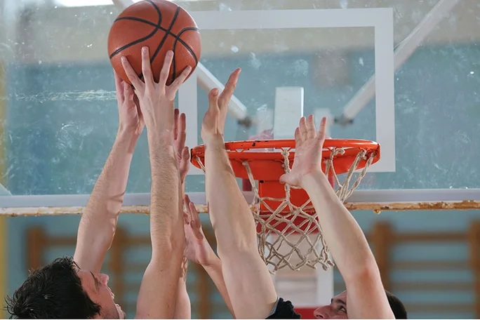 Basketballspiel (Foto: Canva) Im Fokus des Bildes sind die Arme mehrerer Spieler, die versuchen, den Basketball in den Korb zu manövrieren, oder die versuchen, ihn zu blocken.