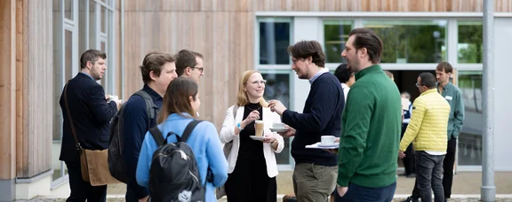 Exchanging ideas and making contacts (Photo: UW/H | Michael Schwettmann) People in conversation. A wooden building façade can be seen in the background.