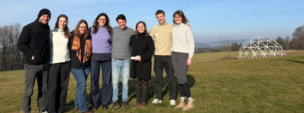 Acht Personen stehen für ein Gruppenfoto auf einer Wiese. Im Hintergrund ist eine kuppelförmige Kunstskulptur zu sehen.