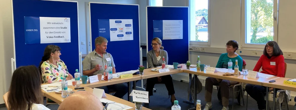 Die Forschergruppe sitzt zusammen an mehreren Tischen vor blauen Pinnwänden.