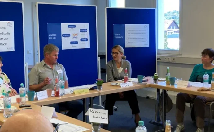 Die Forschergruppe sitzt zusammen an mehreren Tischen vor blauen Pinnwänden.