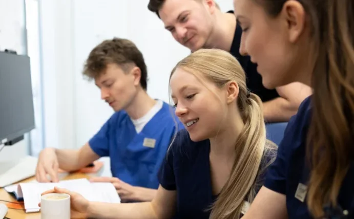Vier Studierende sitzen vor zwei Bildschirmen und schauen auf Dokumente, die vor ihnen liegen.