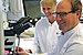 Ein Mann sitzt an einem Mikroskop und lächelt in die Kamera. Hinter ihm ist eine Frau zu sehen.