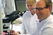 Ein Mann sitzt an einem Mikroskop und lächelt in die Kamera. Hinter ihm ist eine Frau zu sehen.