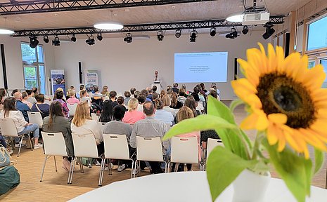 Ein Publikum sitzt in einem Saal und hört einem Speaker zu. Im Vordergrund steht eine Sonnenblume auf einem Stehtisch.