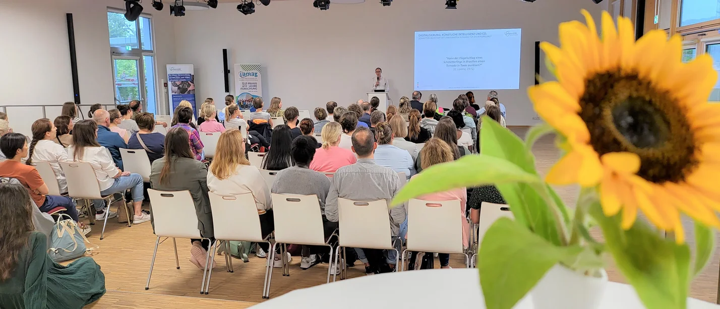Ein Publikum sitzt in einem Saal und hört einem Speaker zu. Im Vordergrund steht eine Sonnenblume auf einem Stehtisch.