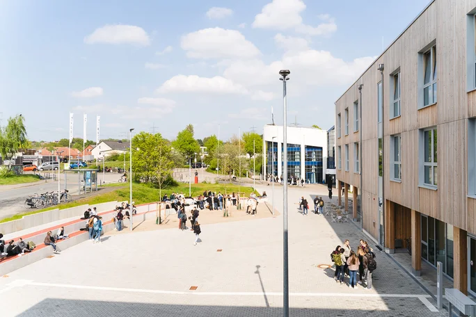 View of the campus with the main and timber buildings (Photo: UW/H | Johannes Buldmann) View over the UW/H campus. You can see the wooden building on the right and the main building in the background. Students walk, stand or sit on the forecourt.