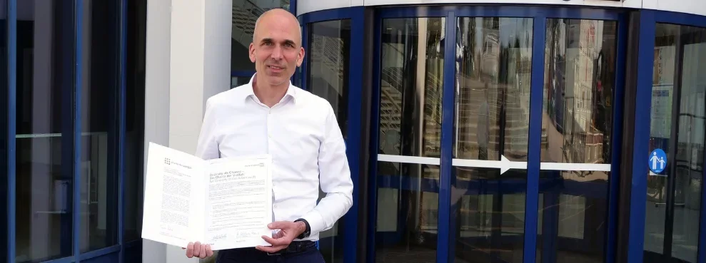 Vizepräsident Dr. Dirk Jakobs steht vor dem Universitätsgebäude und hält eine Urkunde hoch.