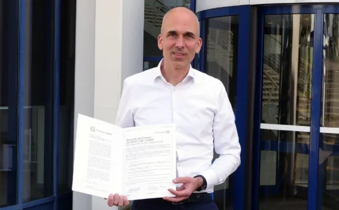 Vizepräsident Dr. Dirk Jakobs steht vor dem Universitätsgebäude und hält eine Urkunde hoch.