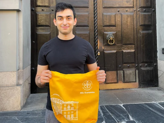 Ein junger Mann steht vor einer großen braunen Tür und hält einen orangenen Beutel in die Kamera.