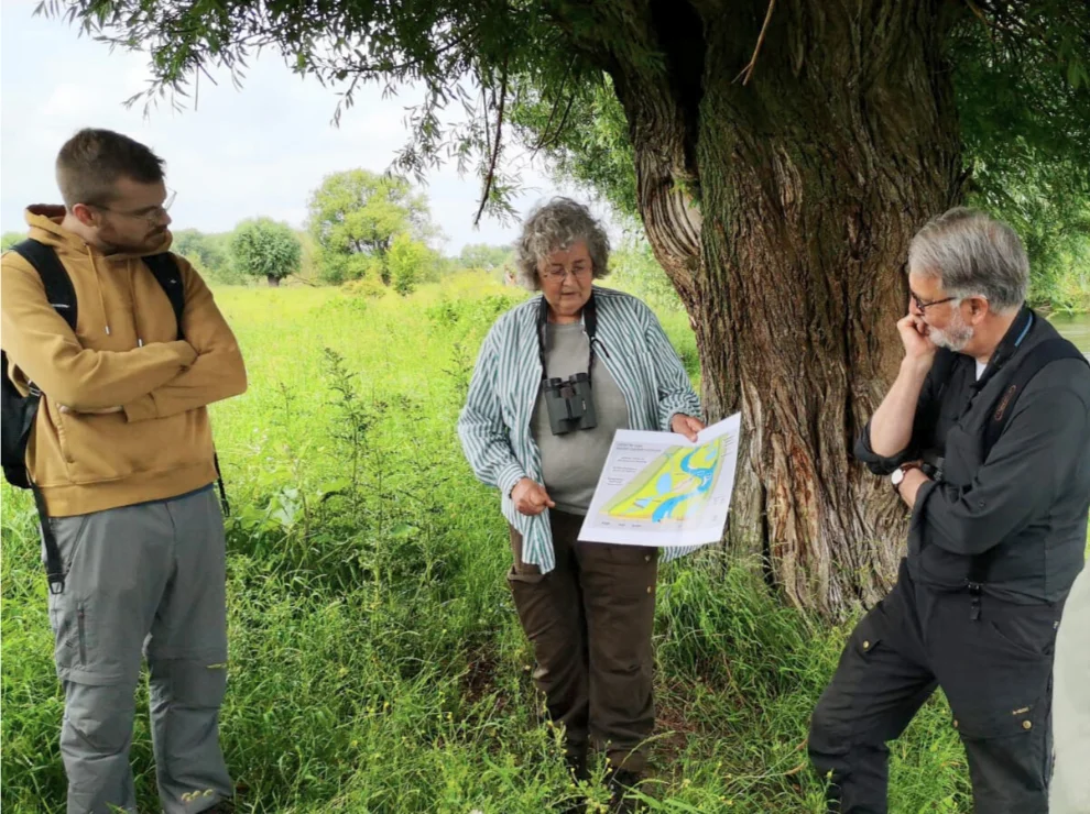 Drei Personen stehen in der Natur und schauen auf eine Karte, die die Frau in der Mitte in der Hand hält.