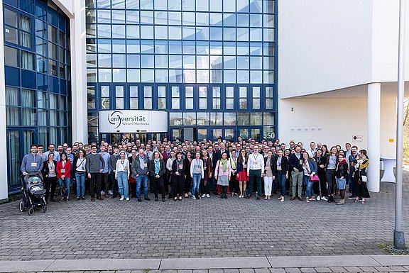 Eine große Personengruppe posiert für ein Gruppenbild. Hinter ihnen ist ein verglastes Gebäude.
