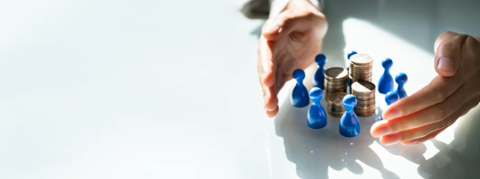 Ein Symboldbild: Zwei Hände umkreisen Spielfiguren und Geldmünzen.