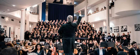 Photo of a conductor standing with his back to the audience. The orchestra and choir are seated behind him.