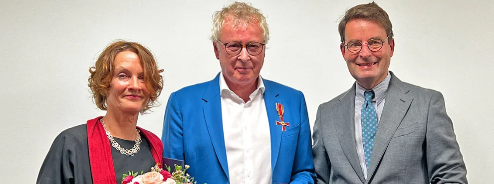 Prof. Dr. Boris Zernikow hat das Bundesverdienstkreuz am Bande verliehen bekommen, er steht zwischen seiner Ehefrau und dem Regierungspräsidenten der Bezirksregierung Münster, Andreas Bothe und lächelt in die Kamera.