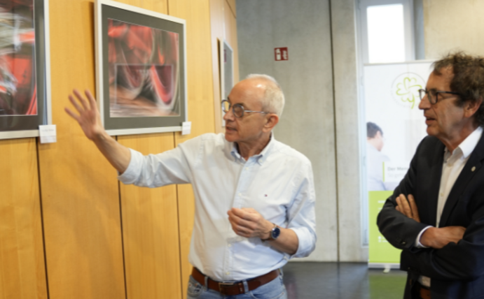 Auf drei Etagen können Patient:innen und Kunstliebhaber:innen in der Zahnklinik Fotografien bestaunen. (Foto: UW/H | Julian Kliem) Ein Mann erklärt einem anderen Mann eine Fotografie.