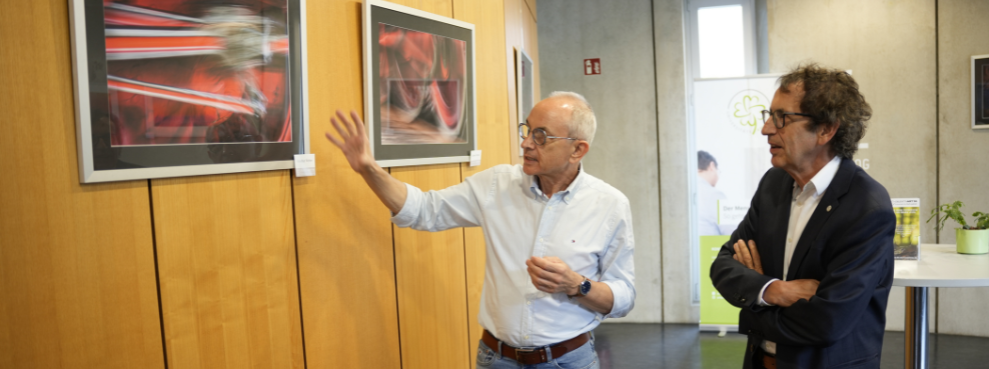 Auf drei Etagen können Patient:innen und Kunstliebhaber:innen in der Zahnklinik Fotografien bestaunen. (Foto: UW/H | Julian Kliem) Ein Mann erklärt einem anderen Mann eine Fotografie.