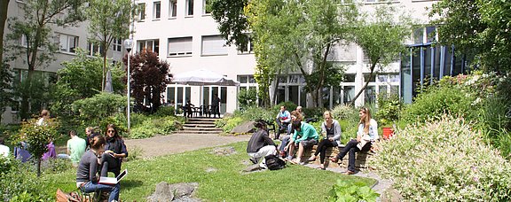 Der Garten ist Dialog- und Begegnungsstätte von Studierenden, Mitarbeitenden und der lokalen Gemeinschaft (Foto: UW/H) Außenbereich vor der Cafeteria im Hauptgebäude mit kleinen Gruppen von Studierenden