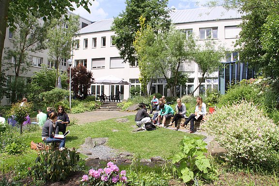 Der Garten ist Dialog- und Begegnungsstätte von Studierenden, Mitarbeitenden und der lokalen Gemeinschaft (Foto: UW/H) Außenbereich vor der Cafeteria im Hauptgebäude mit kleinen Gruppen von Studierenden
