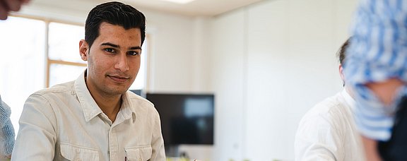 Bewirb dich für den Studiengang Management (M. Sc.) an der UW/H. (Foto: UW/H | Johannes Buldmann) Ein junger Mann sitzt an einem Tisch in einem Seminarraum.