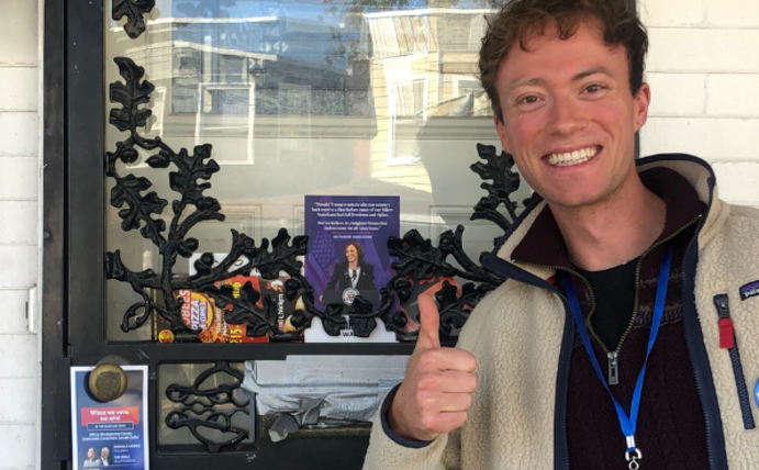 Ein junger Mann steht mit hochgestrecktem Daumen vor einer Haustür und lacht in die Kamera. Er trägt einen blauen Wahlhelfer-Button. Neben im an der Tür kleben Wahlflyer für Präsidentschaftskandidatin Kamala Harris.