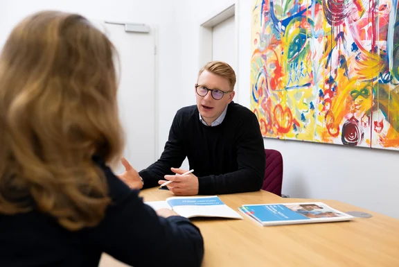 Eine Frau und ein Mann sitzen sich in einem Beratungsgespräch gegenüber. Zwischen ihnen liegen Unterlagen auf dem Tisch.