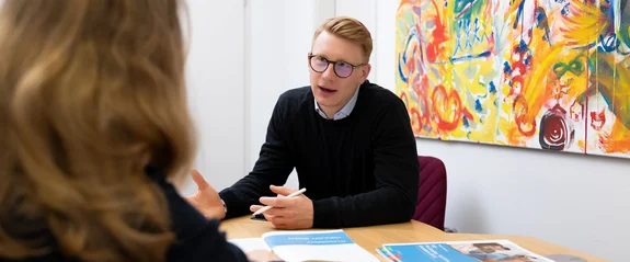 Eine Frau und ein Mann sitzen sich in einem Beratungsgespräch gegenüber. Zwischen ihnen liegen Unterlagen auf dem Tisch.