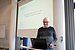Ein Mann steht an einem Rednerpult mit Notebook. Hinter ihm wird eine Präsentation an die Wand projiziert.