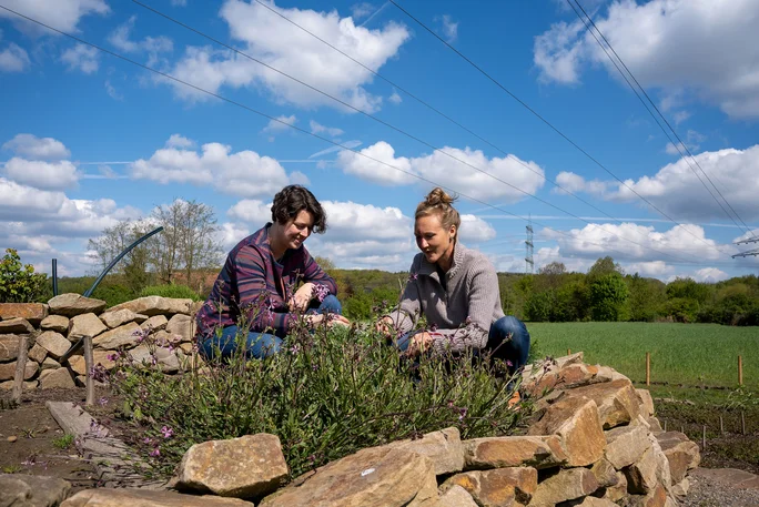 Zwei Frauen knien vor einer gemauerten Kräuterspirale und inspizieren die Pflanzen.