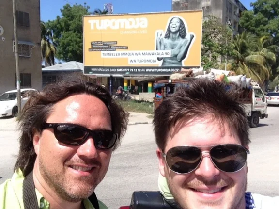 In Tansania hat Oliver Skopec das E-Commerce- und Logistik-Start-up Tupomoja aufgebaut. (Foto: Oliver Skopec) Selfie von zwei Männern vor einer gelben Werbetafel mit der Aufschrift Tupomoja.