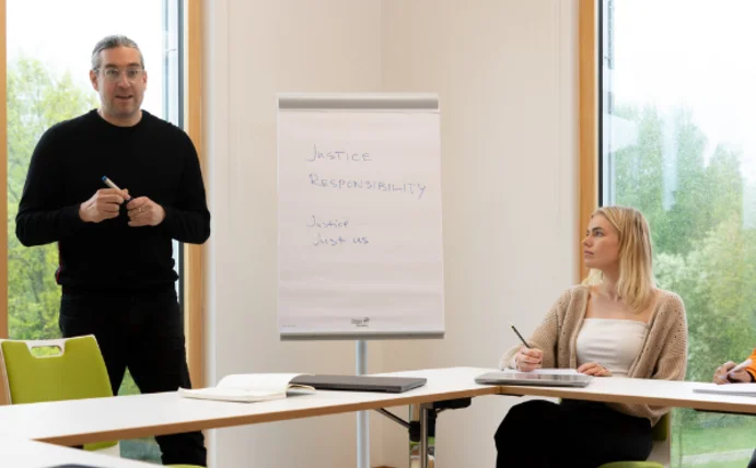 Man sieht Studierende in einem Seminarraum mit einem Dozenten, der zu ihnen spricht.