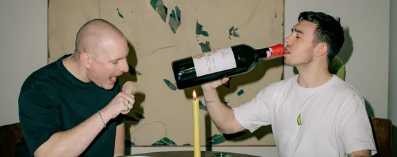 Julian Seitlinger (links) und Adrian Parpat sind die Gründer von Cracky Dining. (Foto: privat) Zwei Männer sitzen an einem Tisch und essen. Einer von ihnen hält sich eine überdimensional große Weinflasche an den Mund.