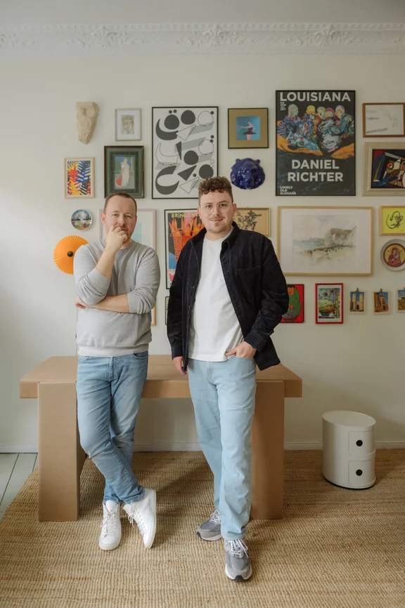 Foto von zwei Männern, die vor einer Bilderwand stehen. Hinter ihnen steht ein Tisch aus Pappe.