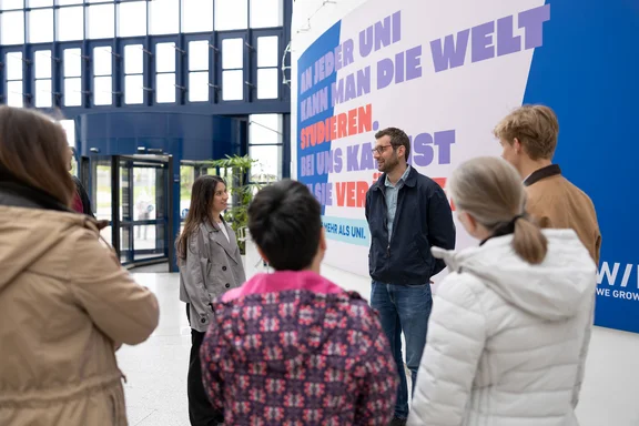 Mach dir mit unseren Infoangeboten ein Bild von der Uni Witten/Herdecke. Eine Gruppe von Personen steht vor einem Plakat.